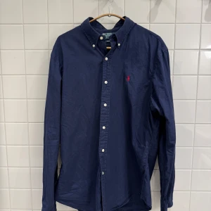 Marinblå skjorta från Ralph Lauren – Storlek L - Superfin marinblå skjorta från Ralph Lauren i storlek L. Skjortan är i superfint skick och har en tidlös design som passar perfekt till både vardag och fest. Kvaliteten är hög och materialet känns mjukt och bekvämt mot huden. Ett klassiskt plagg som ger en stilren look!
