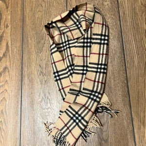 Beige rutig halsduk från Burberry - Säljer en klassisk beige rutig halsduk från Burberry. Den har ett tidlöst mönster med svarta, röda och vita rutor. Halsduken är perfekt för att hålla dig varm och stilren under höst och vinter. Den är i nyskick och har fransar i ändarna för en extra touch av elegans.