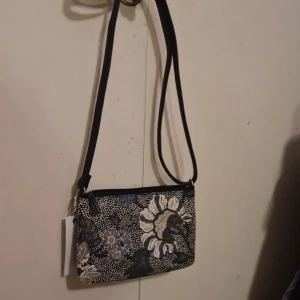 Blommig axelväska från Ceannis - Snygg axelväska från Ceannis med ett blommigt mönster i svart, vitt och blått. Väskan har en justerbar axelrem och en praktisk dragkedja upptill. Perfekt för både vardag och fest!