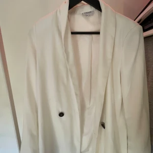 Vit kavaj från Vero Moda - Säljer en stilren vit kavaj från Vero Moda i storlek M. Kavajen har en klassisk design med knappar framtill och är perfekt för både vardag och fest. Den är i mycket bra skick och passar till alla säsonger. Perfekt för att ge din outfit en elegant touch! Knappen på vänster sida har gått sönder