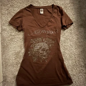 Brun t-shirt med paljetter från Ed Hardy - Säljer en brun t-shirt från Ed Hardy med glittrande paljettdetaljer och ett coolt tryck på framsidan. T-shirten har en v-ringad hals och korta ärmar, den är insydd i midjan för att passa mig så den sitter mer som en S men går lätt att sprätta upp och kan fixa innan ja skickar den😊Han själv inte använt den så mycket men tror den har nåt litet hål nån stans och sen tappade stenar som man kan se på bilden