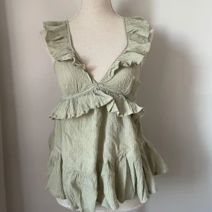 Grön ärmlös topp med volanger - Säljer en söt grön ärmlös topp med volanger. Toppen har en v-ringad design och är perfekt för sommardagar. Materialet är lätt och luftigt, vilket gör den bekväm att bära. Från ASOS, storlek 34 (S). Sparsamt använd - så mycket fint skick :) 💚