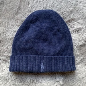 Mörkblå mössa från Polo Ralph Lauren - Säljer en snygg mörkblå mössa från Polo Ralph Lauren. Den är stickad och har en ribbad kant för extra stil. Perfekt för att hålla värmen under kalla dagar. Den ikoniska loggan är broderad framtill. Passar både till vardags och lite finare tillfällen. Med väldigt skönt material! Skriv för fler frågor eller bilder!