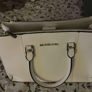 Vit handväska från Michael Kors - Säljer en stilren vit handväska från Michael Kors i mycket bra skick. Väskan har en praktisk dragkedja och eleganta gulddetaljer. Perfekt för både vardag och fest!