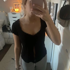Svart topp med spetsdetaljer - Säljer en svart topp med snygga spetsdetaljer längs framsidan. Toppen har korta ärmar och en fin rynkad detalj vid bysten som ger en smickrande passform. Perfekt för både vardag och fest!