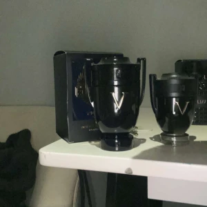 Svart parfymflaska i form av en pokal - Säljer en cool svart parfymflaska som ser ut som en pokal. Den har en glänsande finish och en stilren design med ett V på framsidan. Perfekt för den som vill ha en unik och maskulin doftupplevelse. Flaskan är både snygg och praktisk att ha framme. De är nya och knappt använda om ni vill köpa 1 istället för båda kom dm. Den lilla 400 och den stora 800. Den stora är 100 ml och den lilla är 50ml.