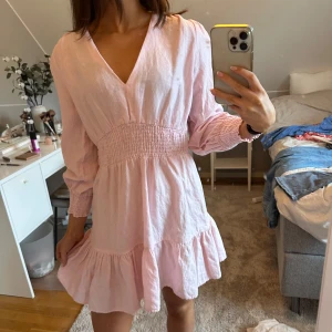 Ljusrosa klänning med volangdetaljer - Säljer en superfin ljusrosa klänning med v-ringning och volangdetaljer. Den har långa ärmar och en smockad midja som ger en snygg passform. Perfekt för vår och sommar!