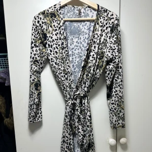 Mönstrad omlottklänning - Snygg och unik omlottklänning med leopardmönster och detaljer i guld. Klänningen har långa ärmar och ett knytband i midjan för en perfekt passform. Perfekt för både vardag och fest!