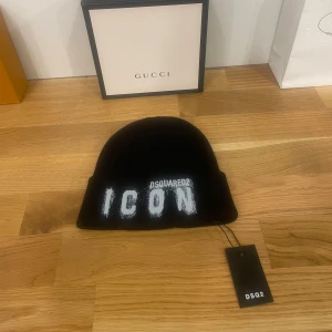 Svart mössa från DSquared2 - Snygg svart mössa från DSquared2 med texten 'ICON' i vitt framtill. Perfekt för att hålla sig varm och stilren under kyliga dagar. Mössan är i nytt skick och har en klassisk passform. Ett måste för modeintresserade!