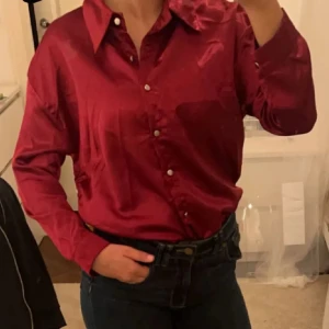 Röd satinblus - Snygg röd satinblus med knappar framtill. Perfekt för festliga tillfällen eller en utekväll. Blusen har en klassisk krage och långa ärmar, vilket ger en elegant look. Passar bra till både jeans och kjol för en stilren outfit.