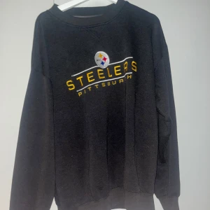 Svart Pittsburgh Steelers sweatshirt - Säljer en svart sweatshirt med Pittsburgh Steelers-logga. Tröjan har långa ärmar och en klassisk rund hals. Perfekt för sportiga tillfällen eller bara för att chilla i. Den är i bra skick och superbekväm!