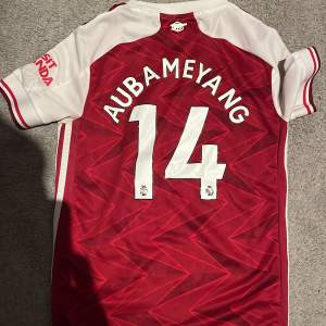 Säljer en snygg Arsenal fotbollströja i rött med vita detaljer. Tröjan har AUBAMEYANG och nummer 14 på ryggen. 