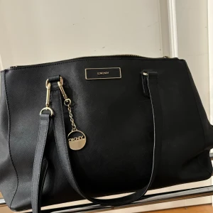 Svart axelväska från DKNY - Snygg svart axelväska från DKNY i skinn med gulddetaljer. Väskan har en praktisk dragkedja och en elegant design som passar perfekt för både vardag och fest. Den har även en dekorativ DKNY-berlock som ger en extra touch av stil.