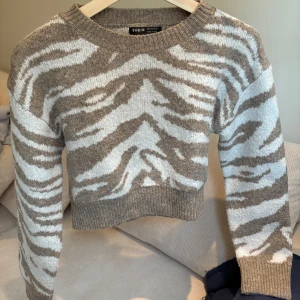 Zebramönstrad stickad tröja - Säljer en zebramönstrad tröja från SHEIN i beige och vit. Den är croppad och har långa ärmar, i storleken S. Materialet är en mix av nylon, polyester och akryl, vilket gör den både mjuk och bekväm. 