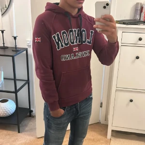 Vinröd hoodie med London-tryck - Säljer en snygg vinröd hoodie med texten 'London England' och Union Jack-flagga på ärmen. Perfekt för höst och vinter med sin mysiga huva och känguruficka. Passar både till vardags och chillkvällar. Hör av dig om du är intresserad! 😊