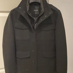 Svart rock från Jack & Jones - Snygg svart rock från Jack & Jones i mycket bra skick. Den har en stilren design med knappar framtill och flera praktiska fickor. Perfekt för höst och vinter, och funkar både till vardags och fest. Rocken har långa ärmar och en klassisk krage. Och är knappt använd storlek S