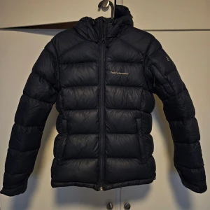 Svart dunjacka från Peak Performance - Säljer en svart dunjacka från Peak Performance i mycket bra skick. Jackan är perfekt för kalla vinterdagar med sin pufferstil och långa ärmar. Den har en praktisk dragkedja och en skön huva för extra värme. Materialet är Pertex Quantum, vilket gör den både lätt och hållbar.