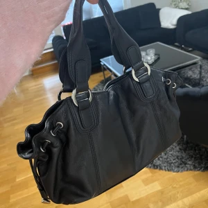 Svart axelväska i skinn - Snygg svart axelväska i skinn med silvriga detaljer. Väskan är i mycket bra skick. 