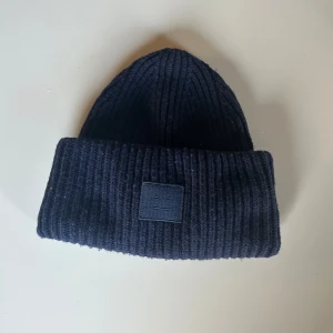 Svart ribbad mössa från Acne Studios - Snygg och stilren svart ribbad mössa från Acne Studios. Perfekt för att hålla värmen under kalla dagar. Den har en enkel design med en diskret logga framtill. Passar till alla outfits och är ett måste för vintern!