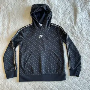 Säljer en svart Nike hoodie med ett diskret mönster av loggor över hela tröjan. Den har en klassisk huva och ribbade muddar. Det finns en grön fläck på ena ärmen annars i bra skick. Perfekt för vardagsbruk eller träning. Storlek 128-137 cm (youth S)