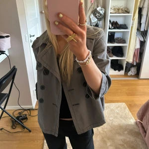 Grå trenchcoat med knappar - Säljer en snygg grå trenchcoat i bra skick. Jackan har stora knappar och trekvartsärmar, perfekt för höst och vår. Den är i en normal passform och passar till både vardag och fest. Ett måste i garderoben för en stilren look!💕💕