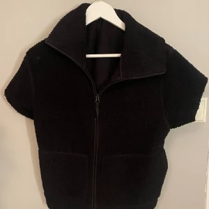 Svart fleeceväst - Säljer en svart fleeceväst i storlek M. Västen har en dragkedja framtill och korta ärmar, perfekt för lager på lager under kyliga dagar. Den är i bra skick och supermjuk att ha på sig. Passar både till vardags och för utomhusaktiviteter.