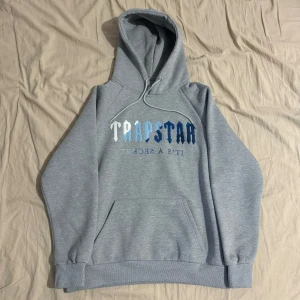 Grå hoodie från Trapstar med blå märke. - Säljer en snygg grå hoodie från Trapstar i storlek M. Den har en cool text på framsidan och är perfekt för både chill och streetwear. Hoodien är i bra skick och superbekväm med en mjuk insida. Perfekt för höst och vinter! Ganska tight fit.