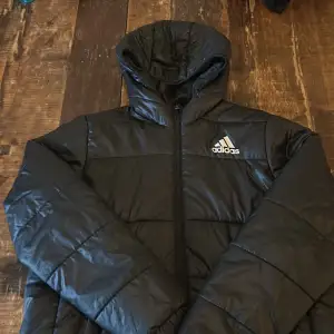 Säljer en svart pufferjacka från Adidas i mycket bra skick. Jackan har en dragkedja framtill och en mysig huva. Perfekt för kyliga dagar och passar både höst och vinter. Den är långärmad och har en stilren design med Adidas-loggan på bröstet. Perfekt för dig som vill hålla stilen i kylan!