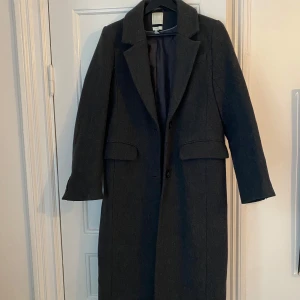 Mörkgrå kappa från H&M ullmix - Säljer en elegant mörkgrå kappa från H&M i ullblandning. Den har en klassisk design med långa ärmar och knäppning fram. Perfekt för höst och vinter, och ger en stilren look. Tillverkad med återvunnen ull från Manteco, vilket gör den både snygg och hållbar.