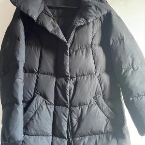 Svart dunjacka - Säljer en svart dunjacka i mycket bra skick. Den är perfekt för vintern med sin långa ärm och quiltade design. Jackan har en dragkedja och knappar för extra skydd mot kylan. Den är både stilren och praktisk, idealisk för kalla dagar.