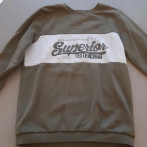 Grön och grå sweatshirt med tryck - Säljer en snygg grön sweatshirt med grått tryck på bröstet där det står 'Superior Best Quality'. Tröjan har långa ärmar och är perfekt för en avslappnad stil. Den är i bra skick och passar perfekt till vardags.