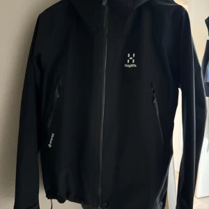 Svart regnjacka från Haglöfs - Säljer en svart regnjacka från Haglöfs i mycket bra skick. Jackan har en praktisk huva och dragkedja framtill. Perfekt för regniga dagar och utomhusaktiviteter. Den är både stilren och funktionell, vilket gör den till ett måste i garderoben. Passar perfekt för höst och vår!
