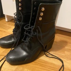 Svarta boots från Tiger of Sweden - Snygga svarta boots i skinn från Tiger of Sweden. De har en rejäl klack och snörning framtill med silverfärgade knappar som detaljer. Perfekta för höst och vinter, och ger en cool touch till vilken outfit som helst. Använd dem till både vardags och fest!