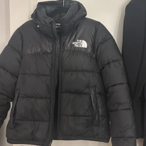 Svart pufferjacka från The North Face - Säljer en svart pufferjacka från The North Face i mycket bra skick. Den är perfekt för kalla vinterdagar med sin tjocka vaddering och har en praktisk dragkedja framtill. Jackan har en huva och långa ärmar, vilket gör den extra mysig och varm. Passar både till vardags och utomhusaktiviteter. Huvan är kan man dessutom få bort. Ordinarie pris ligger på minst 2700kr