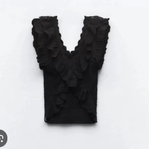 Svart spetsblus med volanger - Snygg svart blus i spets med volanger framtill. Perfekt för en festlig kväll eller en stilfull dag på stan. Den har en elegant design med djup v-ringning och är ärmlös💕