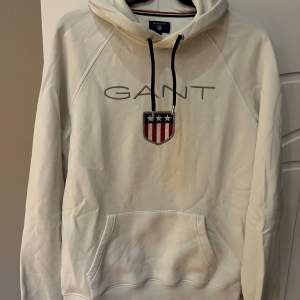 Säljer en snygg vit hoodie från GANT i storlek M. Den har en klassisk design med GANT-loggan och ett coolt emblem på bröstet. Perfekt för en avslappnad stil med känguruficka och justerbar huva. Passar perfekt till både höst och vår!