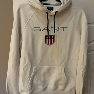 Vit hoodie från GANT - Säljer en snygg vit hoodie från GANT i storlek M. Den har en klassisk design med GANT-loggan och ett coolt emblem på bröstet. Perfekt för en avslappnad stil med känguruficka och justerbar huva. Passar perfekt till både höst och vår!