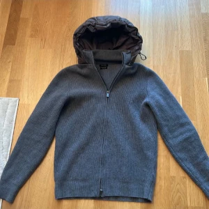 Grå stickad jacka från Massimo Dutti - Säljer en snygg grå stickad jacka från Massimo Dutti i mycket bra skick. Den har en praktisk dragkedja och en quiltad huva som ger extra värme. Perfekt för höst och vår när det är lite kyligt ute. Passar bra till både vardag och lite finare tillfällen.