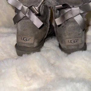 Grå UGG boots med rosett - Superfina grå UGG boots i mocka med fluffigt foder och söta rosetter bak. Perfekta för att hålla fötterna varma och mysiga under kalla dagar. Storlek 37. 