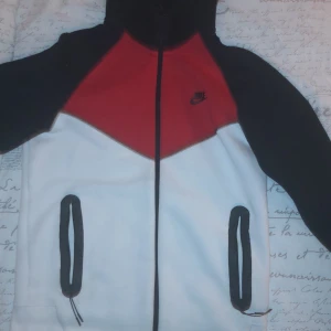 Nike fleecejacka med huva - Säljer en snygg fleecejacka från Nike i rött, svart och vitt. Den har en dragkedja framtill och två fickor med svarta detaljer. Perfekt för höst och vår, med en skön huva för extra värme. Jackan är i mycket bra skick och passar perfekt för både träning och vardag.