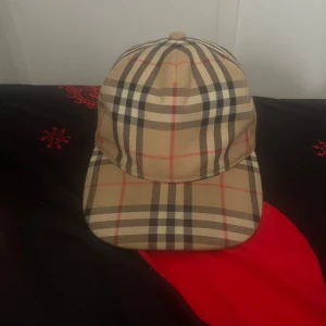 Rutig keps från Burberry - Säljer en snygg rutig keps från Burberry i klassiskt beige, svart och rött mönster. Kepsen har en justerbar rem baktill och är i mycket bra skick. Perfekt för både vardag och festliga tillfällen. Passar de flesta storleken M.Byte hade också funkat bara å skriva för mer info 