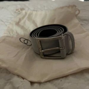 Grått mocka bälte från OneWear - Säljer ett stilrent grått bälte från OneWear i mocka. Bältet har ett klassiskt silverfärgat spänne och är perfekt för både vardag och fest. Det är i mycket bra skick och kommer i sin originalpåse. Det är ett limited edition bälte som aldrig kommer säljas igen. Bältet är 90cm långt