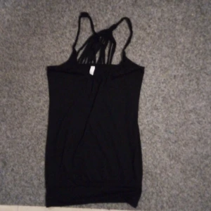 Svart ärmlös topp från Madonna - Snygg svart ärmlös topp från Madonna med smala flätade axelband och strimlad detalj i bak en stilren design. Perfekt för en utekväll eller sommarens fester. Toppen är i bra skick och har en bekväm passform.