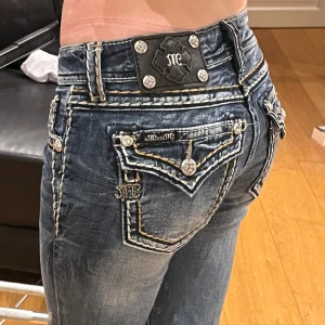 Blå jeans med detaljer - Snygga blå jeans från Miss Me med coola detaljer och broderier. De har låg midja och en tight passform som framhäver figuren. Perfekta för en casual look eller en utekväll. Dekorativa knappar och sömmar ger en unik stil. Midja: 37cm, Innerben: 79cm