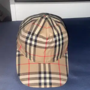 Snygg rutig keps från Burberry i klassiskt beige, svart och rött mönster. Perfekt för att ge din outfit en stilren touch. Justerbar passform baktill med en svart rem. Passar både till vardags och festliga tillfällen.