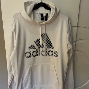 Vit hoodie från Adidas - Säljer en snygg vit hoodie från Adidas i storlek M. Den har en stor logga på framsidan och en klassisk känguruficka. Perfekt för både vardag och träning. Huvan har dragsko för justerbar passform. Superbekväm och stilren! Knappt använd. 