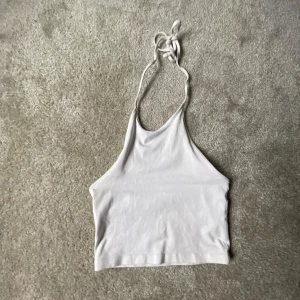 Vit halterneck linne - Säljer ett stilrent vitt halterneck linne i mjukt material. Perfekt för sommardagar eller en avslappnad fest. Linnet är ärmlöst och har en knytning i nacken för justerbar passform. Superbekvämt och lätt att matcha med olika outfits!