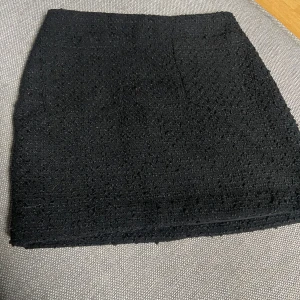 Svart kjol i texturerat tyg - Säljer en svart kjol i texturerat tyg. Den har en enkel och stilren design, perfekt för både vardag och fest.  Passar bra till olika toppar och skor för en chic look. Tyvärr står det inget märke. Det står heller ingen stolkek på men det är väldigt liten i stolek, skulle säga att det är xxs eller xs.