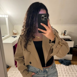 Beige croppad trenchcoat - Säljer en superfin beige croppad trenchcoat med knappar och bälte i ärmsluten. Perfekt för höst och vår, och ger en stilren look till vilken outfit som helst. Jackan har en loose passform och är i mycket bra skick. Passar perfekt till både jeans och klänning!