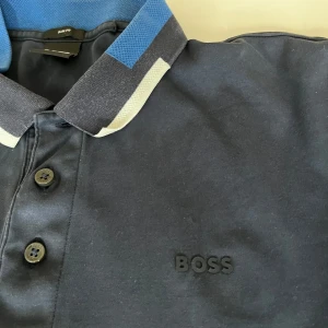Mörkblå pikétröja från BOSS - Säljer en stilren mörkblå pikétröja från BOSS. Den har en snygg krage med blå och vita detaljer samt knappar framtill. Perfekt för både vardag och festliga tillfällen. Den är passar för någon som är kortare och bredare. Är M men passar S.